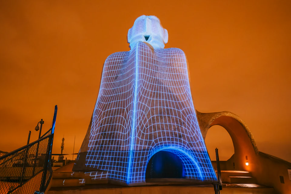A stone sculpture is illuminated with blue grid patterns against an orange sky, featuring arched openings and angular shapes.