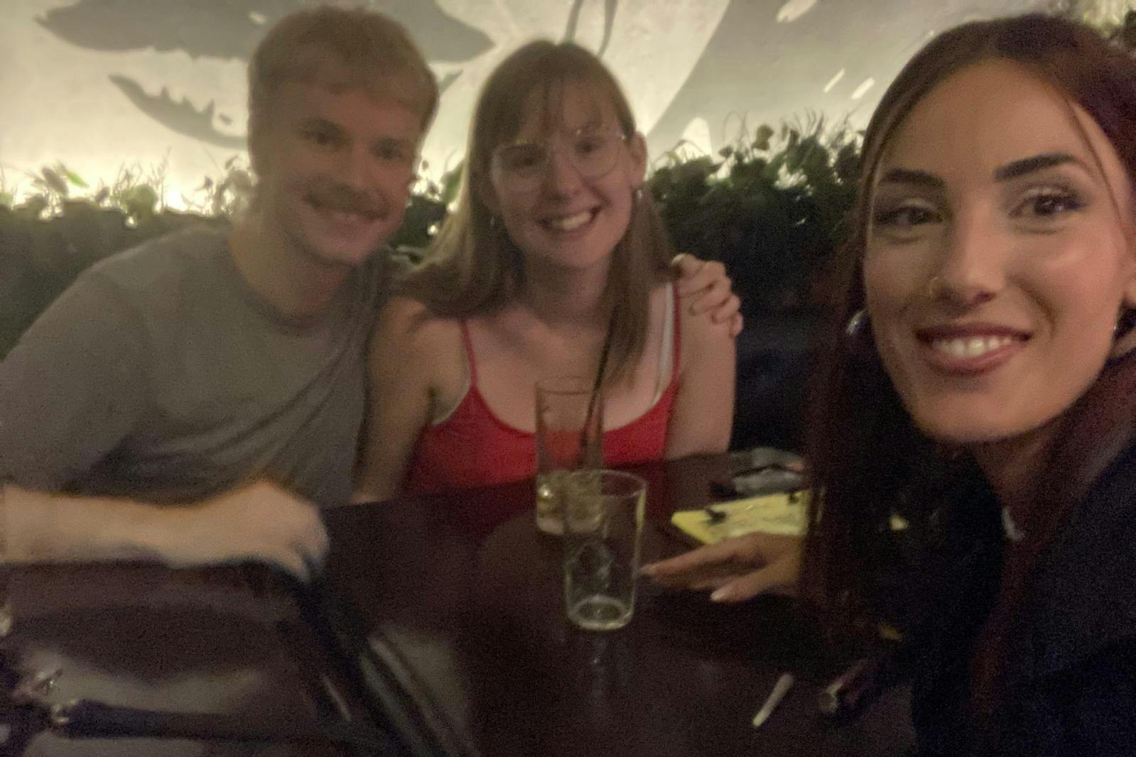 Three people are sitting at a table in a dimly lit setting, smiling at the camera, with drinks and papers on the table.