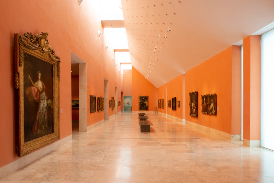 An art gallery with orange walls, framed paintings, a high ceiling, and a marble floor. Benches are placed in the center walkway.