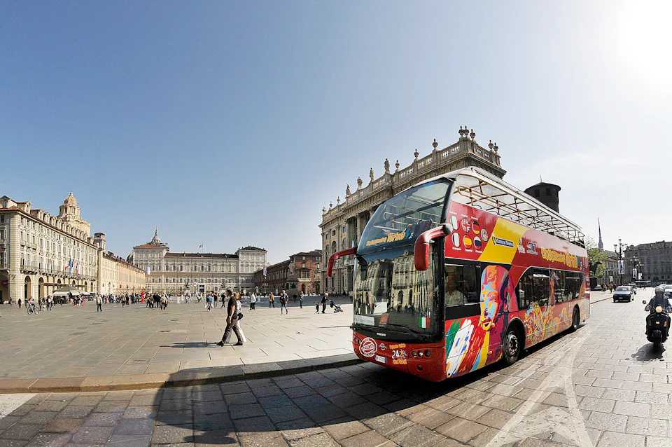 A red double-decker tour bus with colorful graphics parked in a large, historic plaza with people walking and classic buildings in the background.