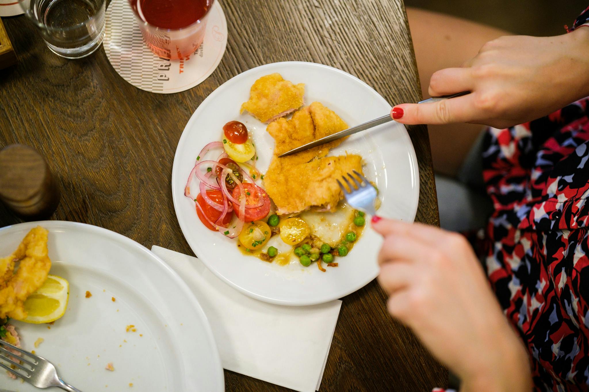 Člověk jí smažené ryby s hráškem, rajčaty, cibulí a citronem. Na dřevěném stole je také vidět nápoj a prázdný talíř.