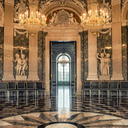 Elegant dekorerad, ljuskrona-belyst hall med marmorpelare, utsmyckade statyer, speglar, svartvitt rutigt golv och stolar.