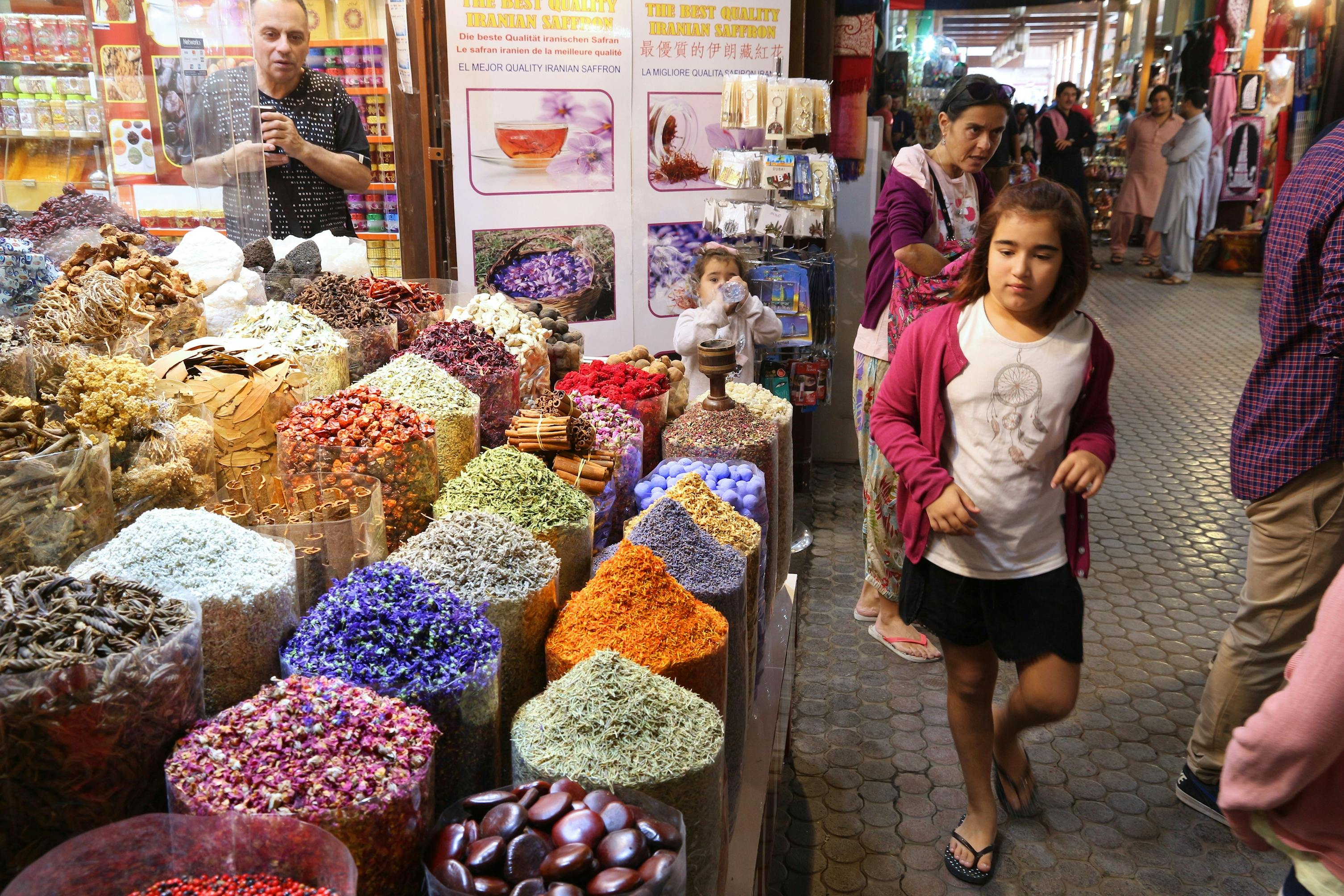 Una bancarella del mercato al coperto espone varie spezie colorate e prodotti secchi, con i clienti, tra cui un uomo che fotografa e bambini che curiosano.