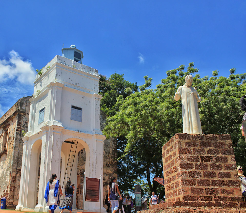 People walking near a tall white bell tower and a statue on a stone pedestal, with trees and a blue sky in the background.