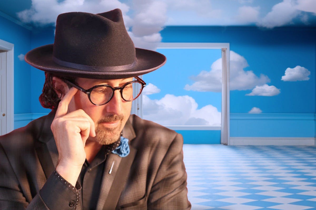 En mand iført briller og en sort hat i et rum med blå vægge og et himmelmaleri med skyer.