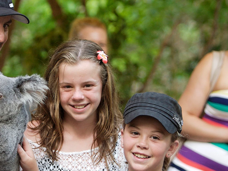 Two girls pet a koala held by a man in uniform with a woman in the background. Trees and greenery are visible.