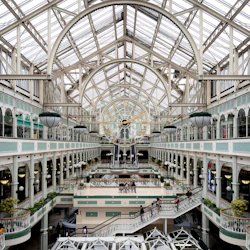 Elegant inomhusgalleria med en stor klocka, välvt glastak, flera våningar, balkonger och dekorativa räcken.