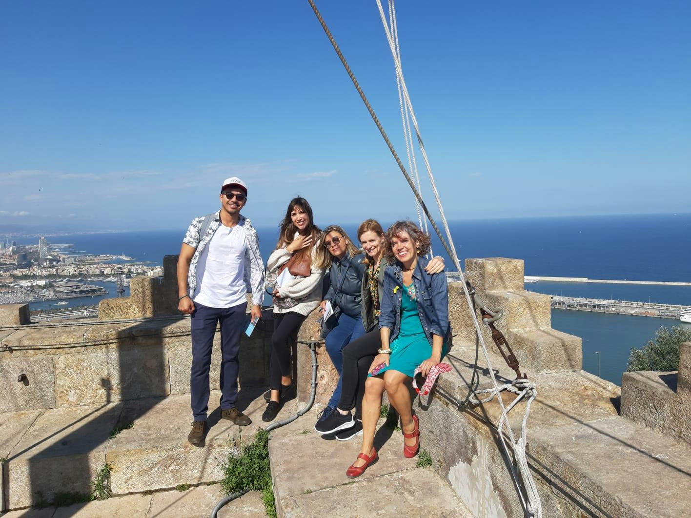 Five people smile and pose on a stone structure with a coastal city and ocean in the background on a clear day.