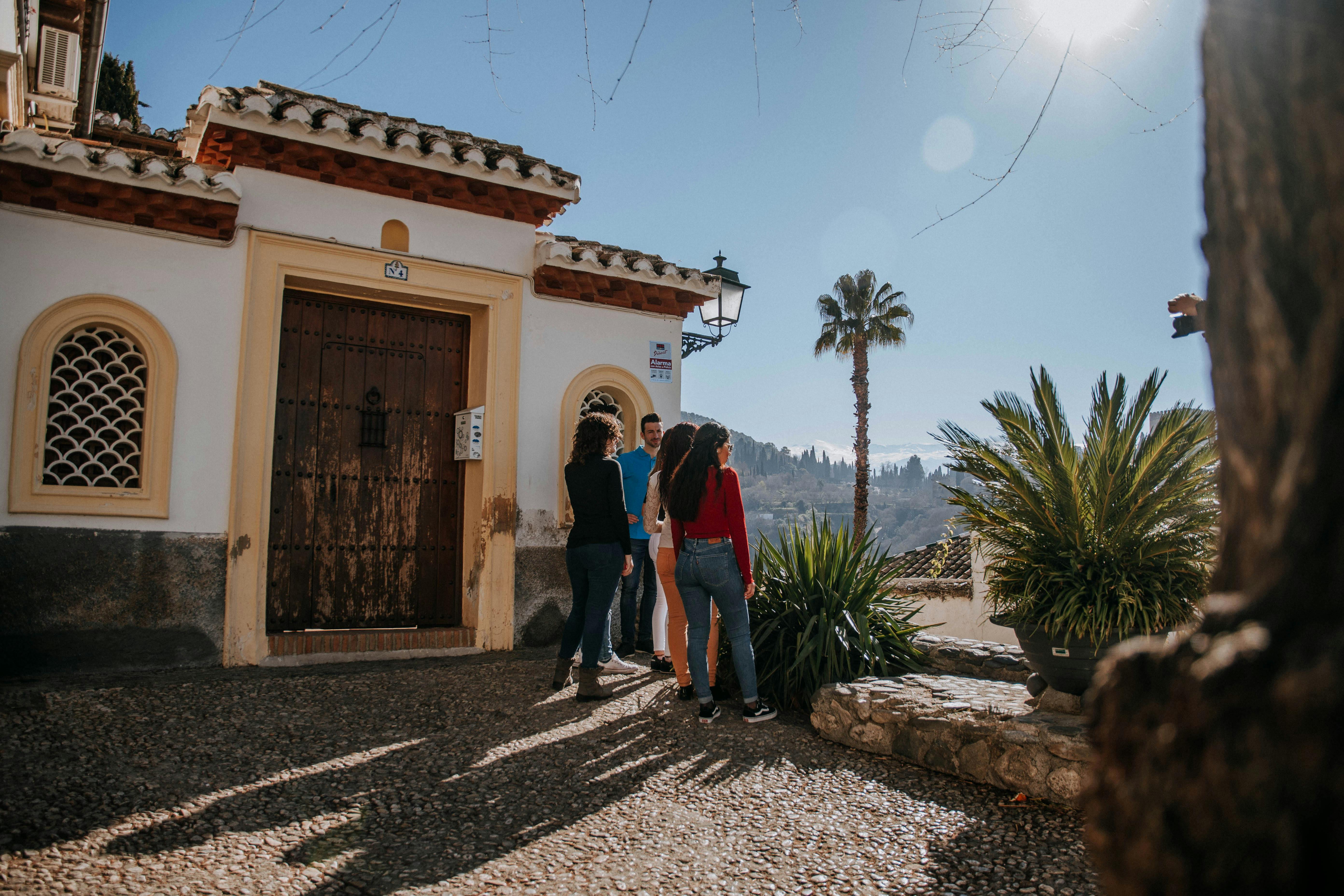 A group of people stand near a wooden door of a white building with arched windows, next to a palm tree and greenery under the sun.