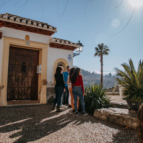 En grupp människor står nära en trädörr i en vit byggnad med välvda fönster, bredvid en palm och grönska under solen.