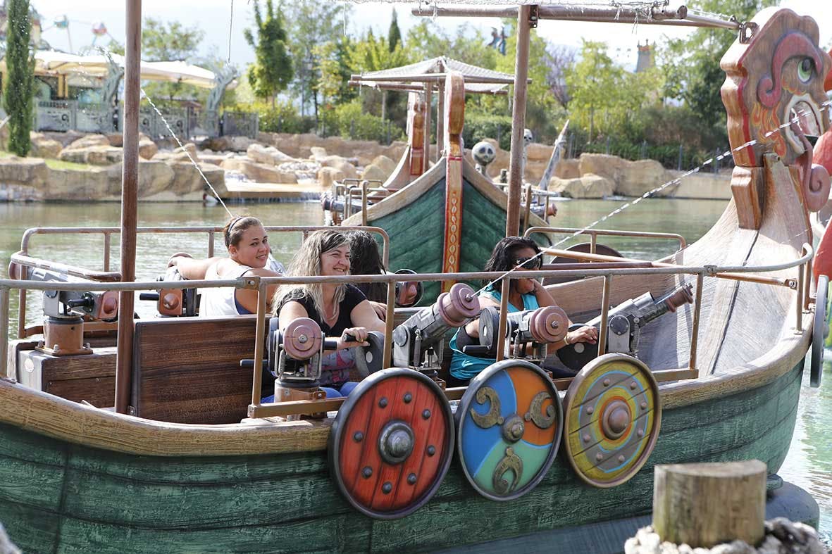 Οι άνθρωποι απολαμβάνουν μια βόλτα με ένα πειρατικό σκάφος σε ένα πάρκο ψυχαγωγίας, με μια λίμνη και πράσινο στο παρασκήνιο.