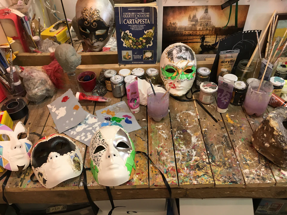 A cluttered artist's workspace with decorated masks, paints, brushes, and an instructional book on cartapesta mask-making.