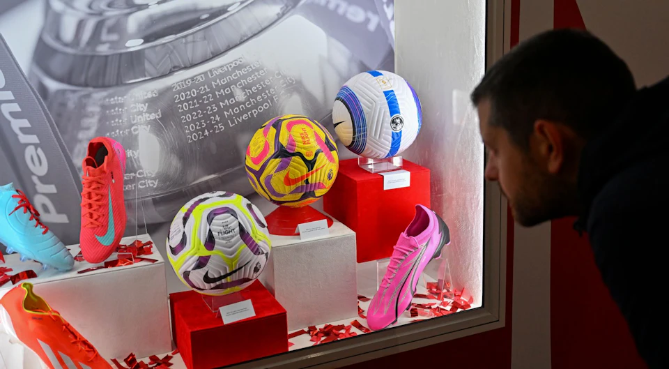 Three soccer balls and a pink soccer shoe displayed on red pedestals. A man is looking at the exhibit. Text on the background wall.