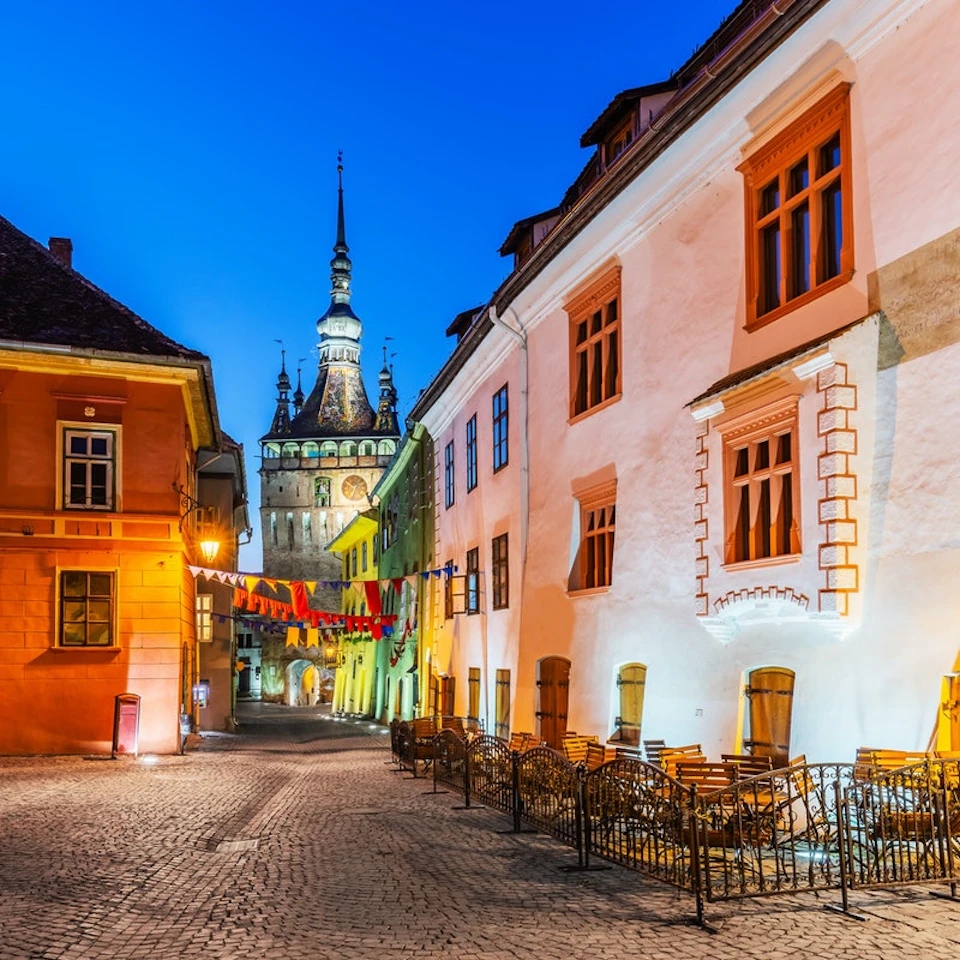 Colorful buildings with illuminated windows line a cobblestone street, with a tower in the background under the evening sky.