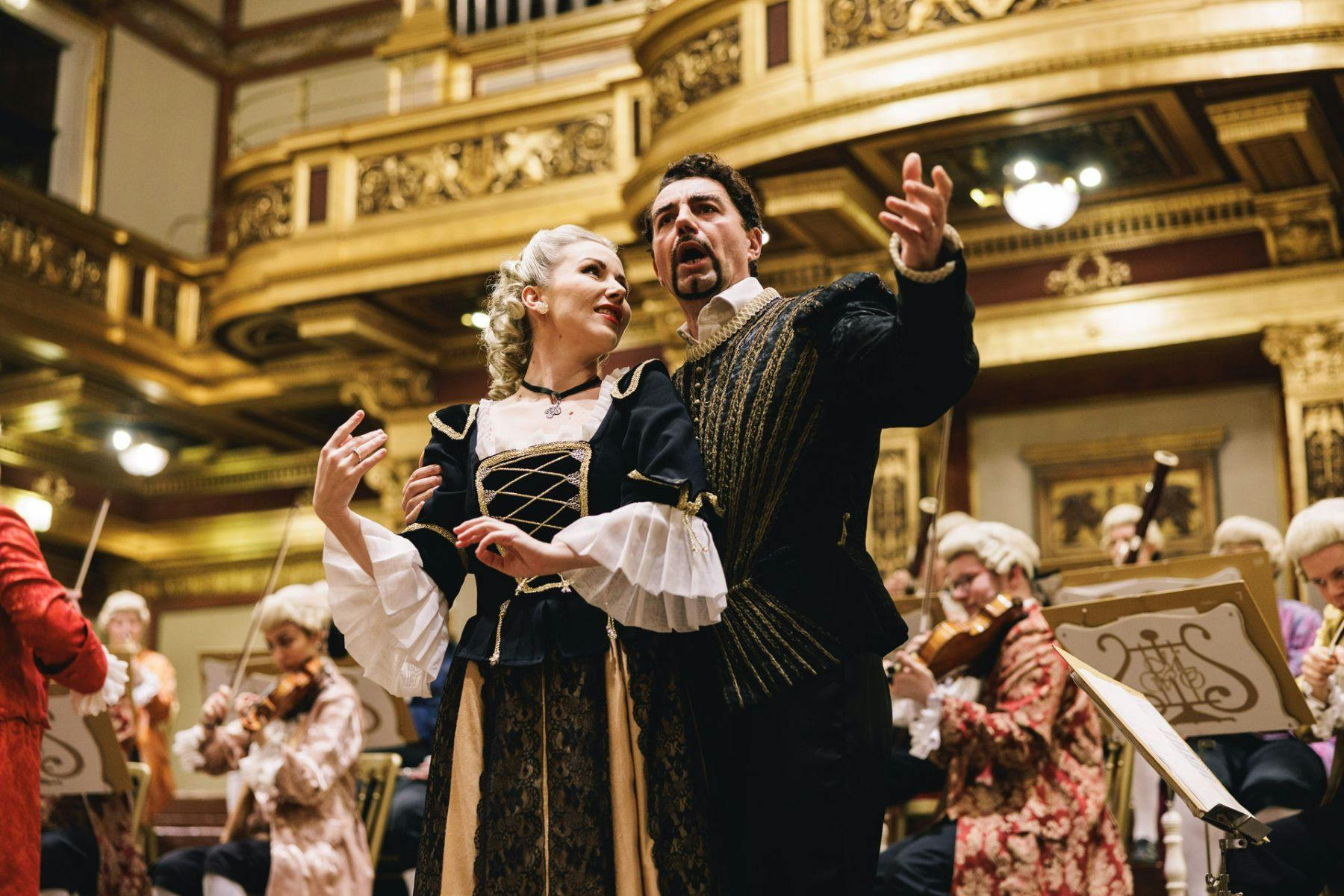 Two opera singers in period costumes perform passionately on stage, with a baroque orchestra playing in the background.