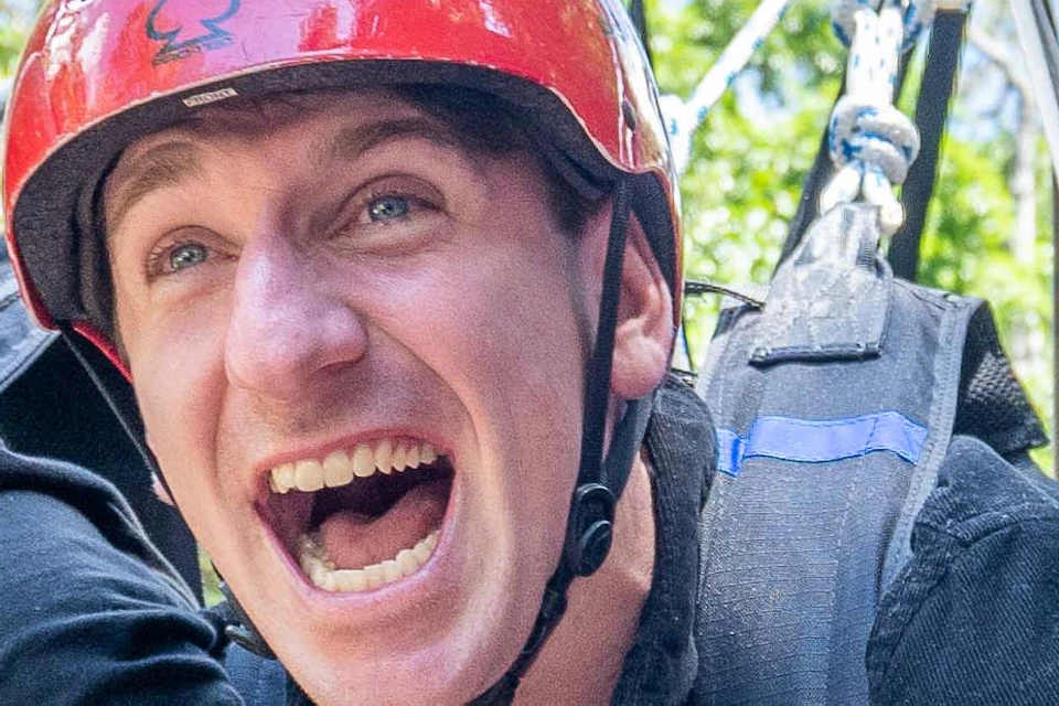 Three people wearing helmets and harnesses enjoy an outdoor zip-lining adventure surrounded by lush trees and a blue sky.