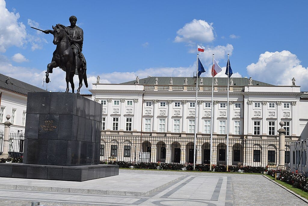 Estàtua ecuestre davant d'un edifici neoclàssic amb tres banderes en pals i un cel brillant i ennuvolat de fons.