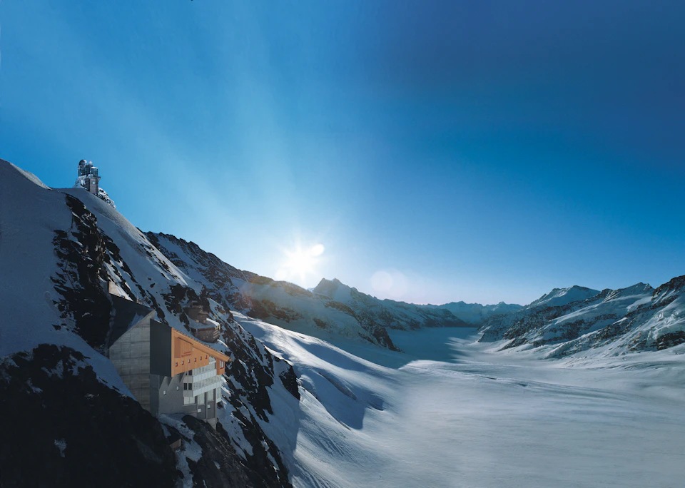 A building situated on a snowy mountainside with a vast snow-covered landscape extending under a clear blue sky and a setting sun.