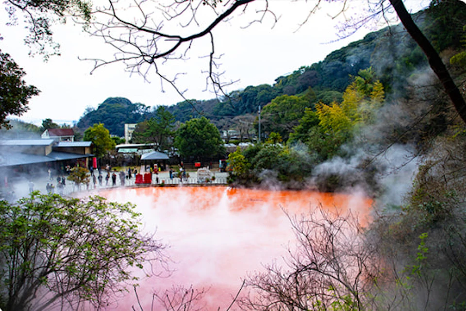 A scenic view of a red-colored hot spring surrounded by trees and buildings, with steam rising, and people nearby.
