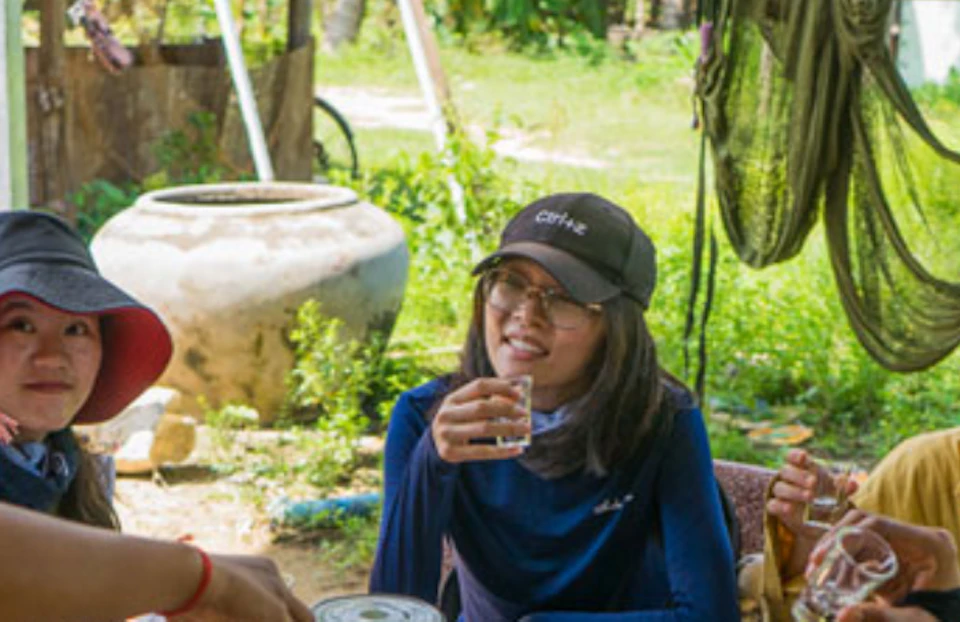 Group of people sitting around a round outdoor table, drinking from glasses, with a water jug and a jar of snacks in the center.