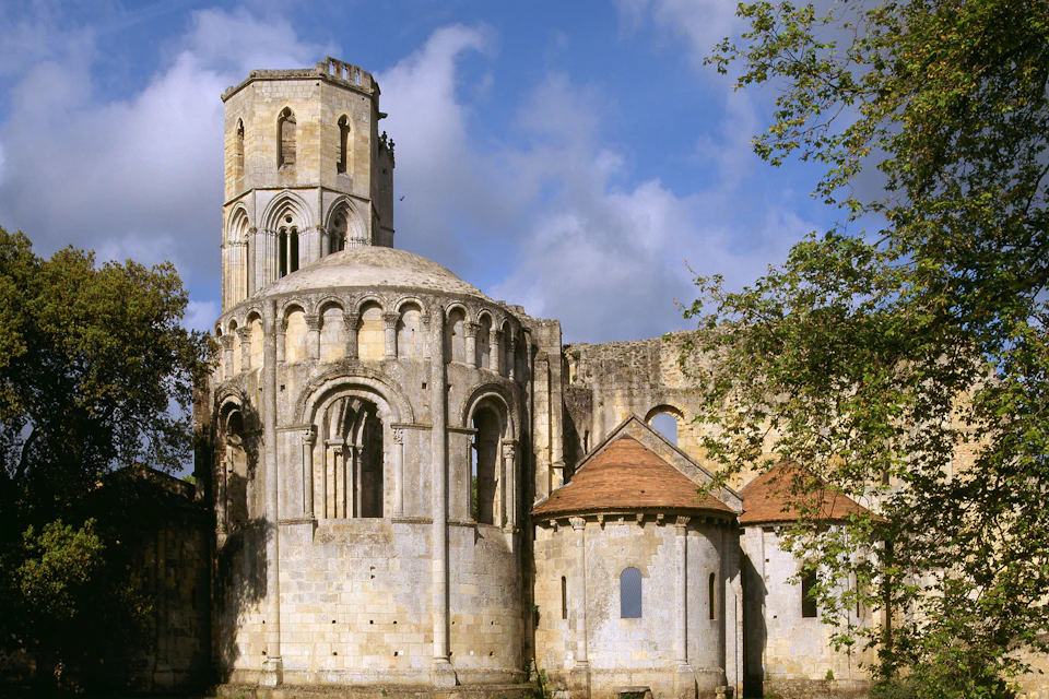 Ancient stone building with arched windows and a cylindrical tower, surrounded by trees under a partly cloudy sky.