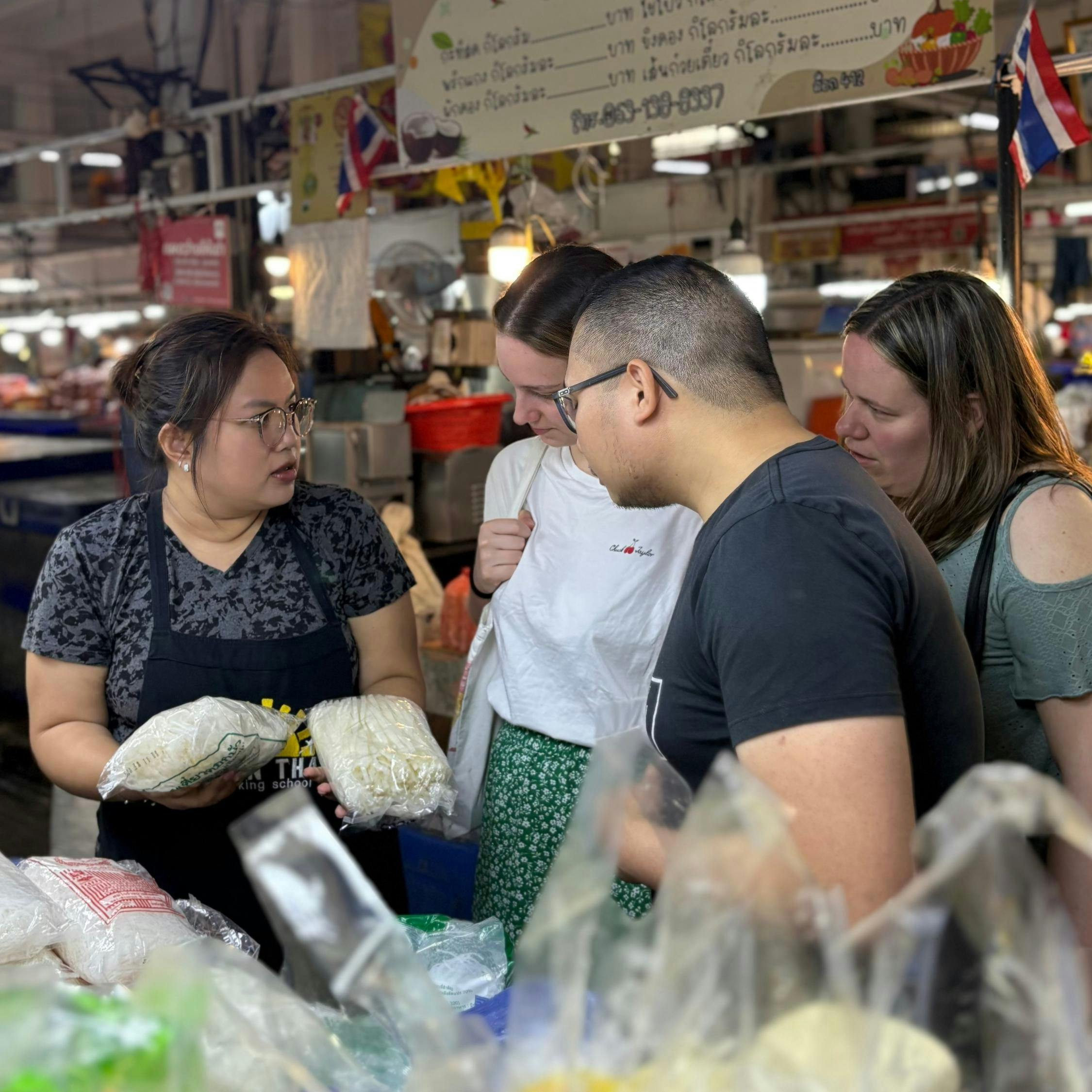 A vendor at a market showing bags of food to a group of three people, with shelves and products visible in the background.