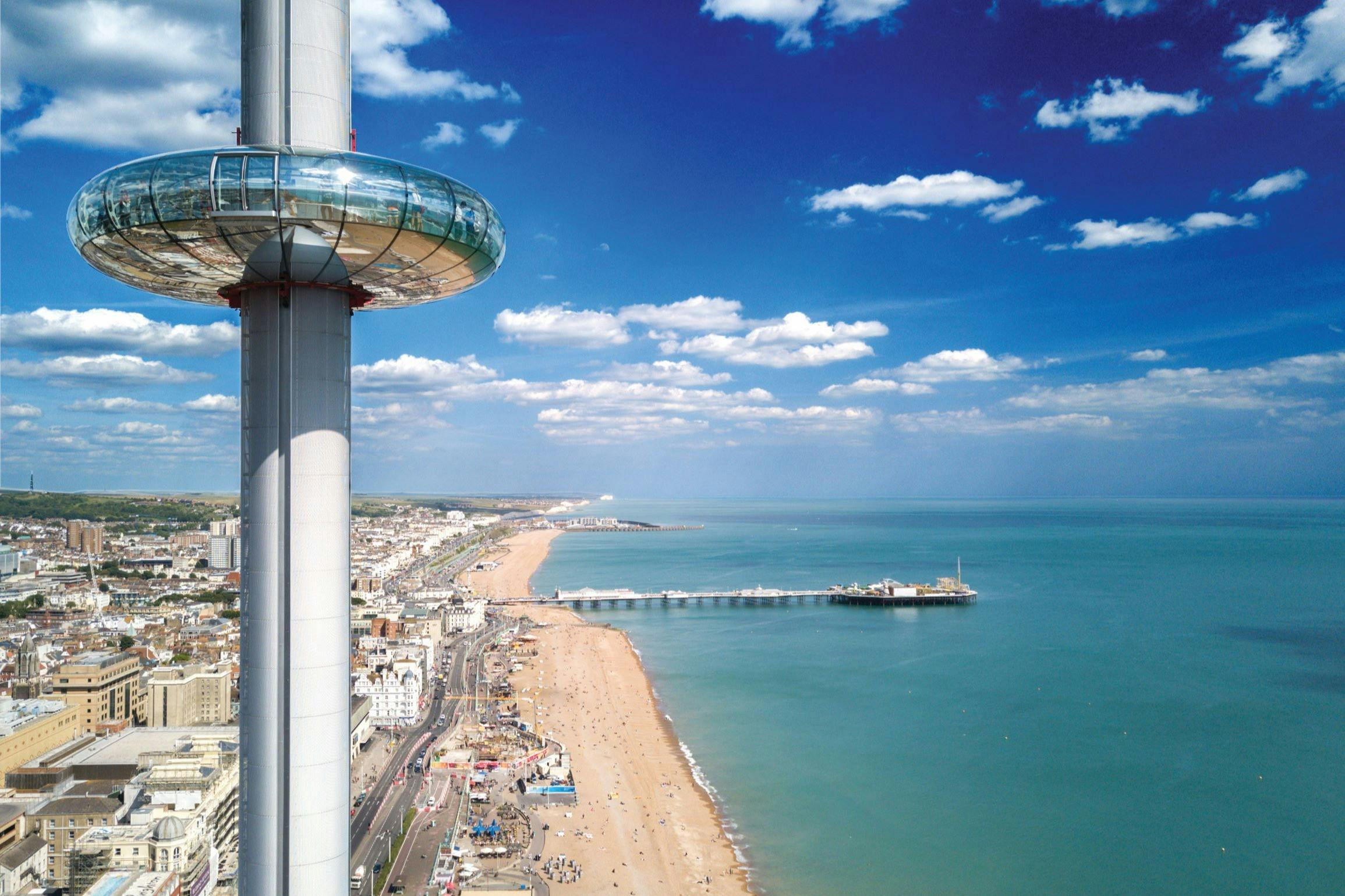 Aerial view of a beach with a pier extending into the ocean, a coastal city, and a tall observation tower. The sky is partly cloudy.