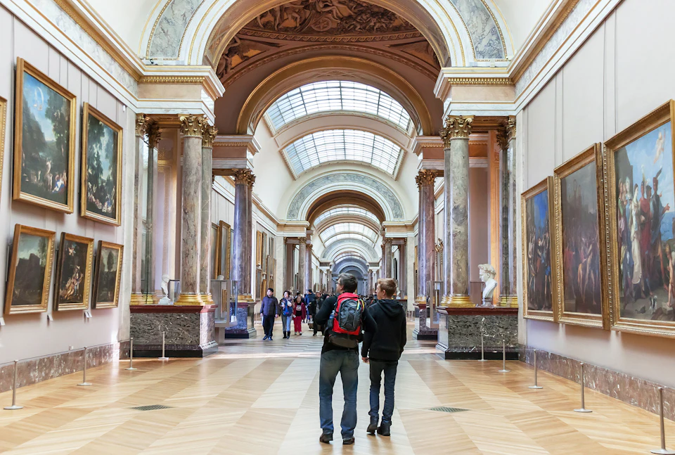 People walking through a spacious art gallery with high arched ceilings and numerous framed paintings on the walls.