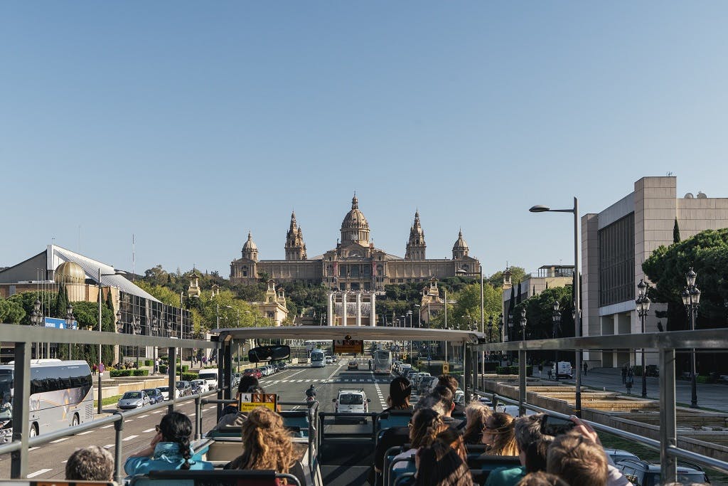Le persone a bordo di un autobus turistico aperto ammirano un grande edificio storico con cupole, percorrendo un&#39;ampia strada cittadina sotto un cielo azzurro e limpido.