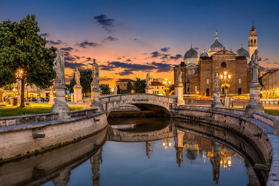 A scenic view of a canal with statues on its sides, a bridge, and a historic building at sunset.