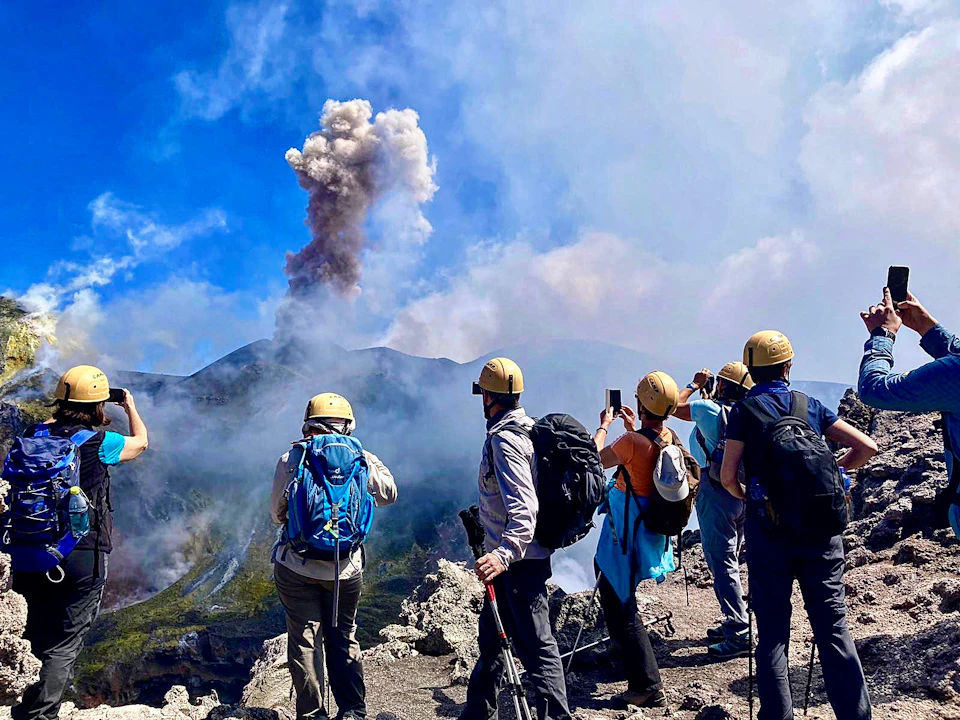 People in hard hats and backpacks observing a volcanic eruption, with smoke rising against a blue sky.