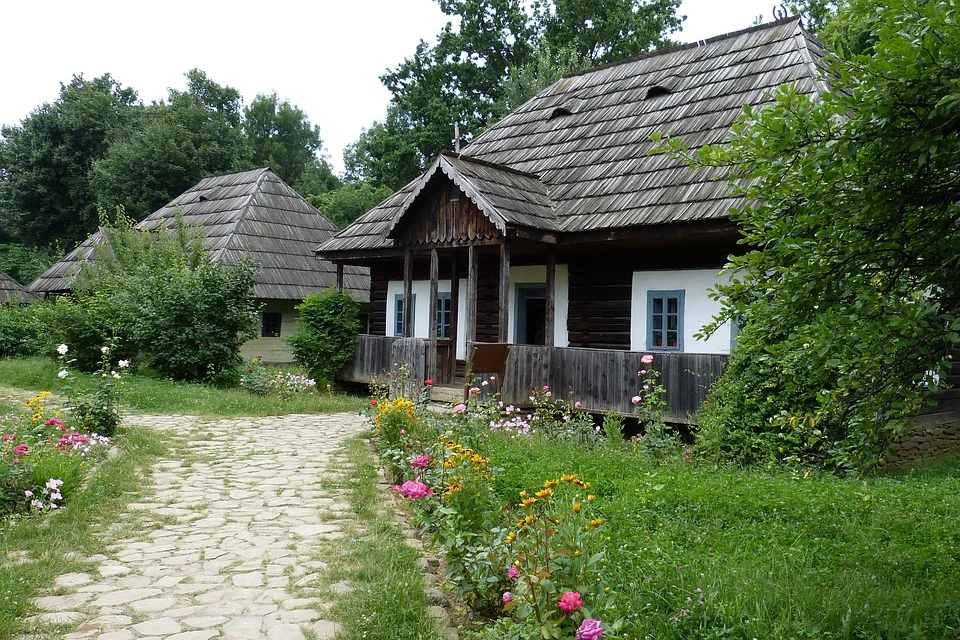 Two rustic wooden houses with shingled roofs and porches, surrounded by lush greenery and colorful flowers along a stone path.