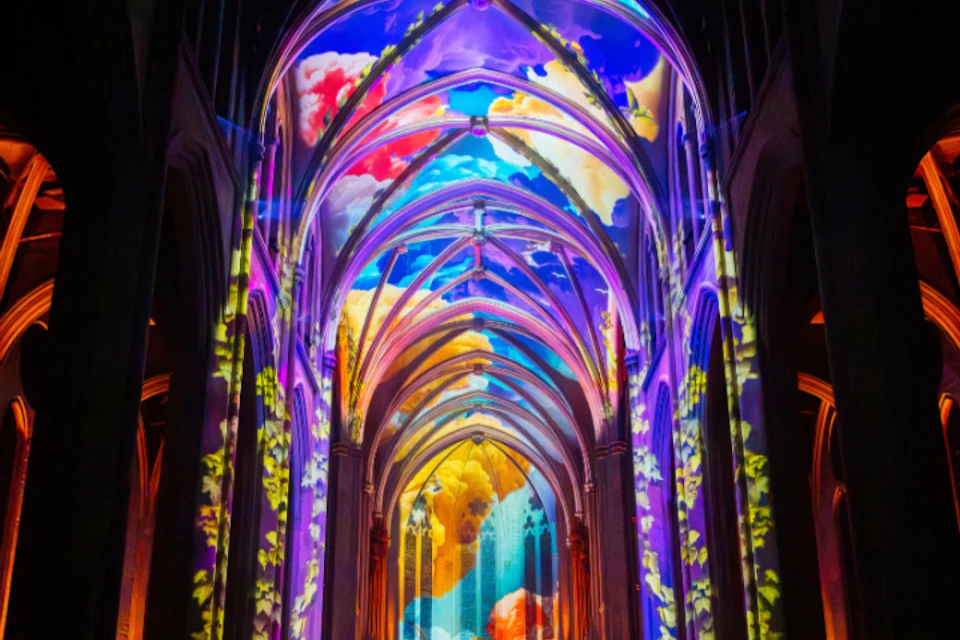 A cathedral interior lit with vibrant, colorful projections of clouds and abstract patterns on the arches and ceiling.