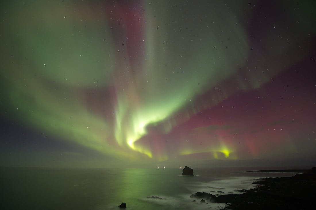 Coloratissime aurore boreali illuminano il cielo notturno sopra un oceano scuro con formazioni rocciose lungo la costa.