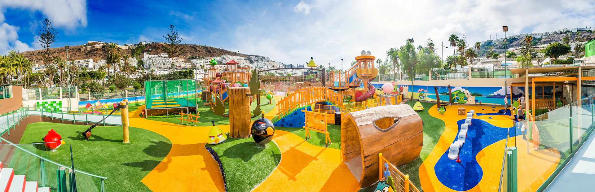 Colourful playground with slides, bridges, climbing structures, and character-themed designs on a sunny day. Apartments visible in the background.