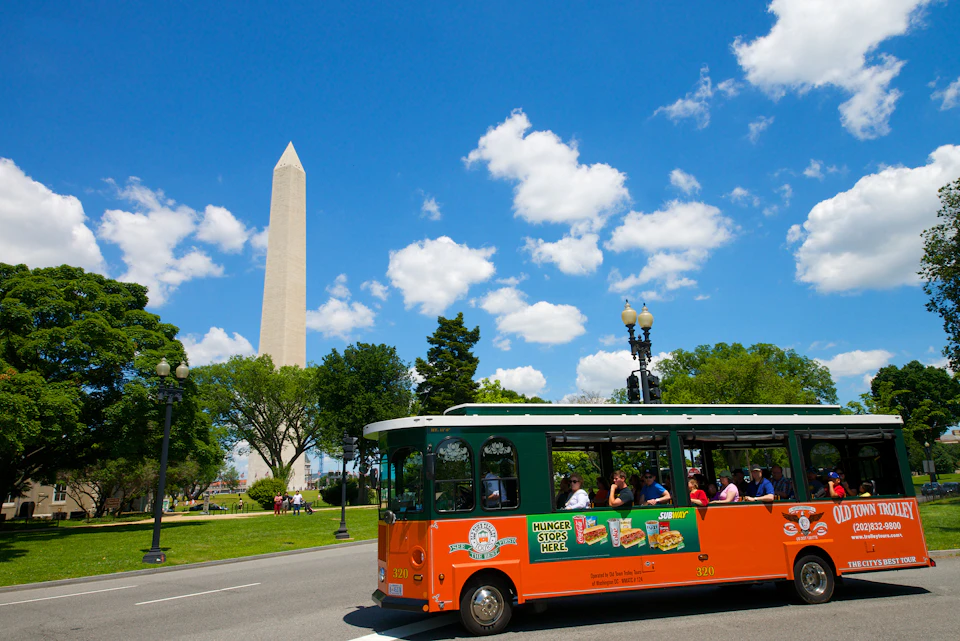 Tour bus driving past the Washington Monument on a clear day with a blue sky and scattered clouds.
