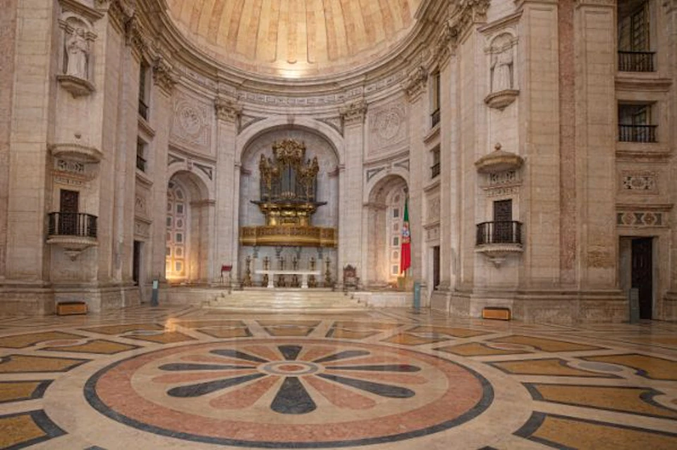 Interior of a grand, ornate cathedral with a large pipe organ at the front, marble columns, intricate wall carvings, and a tiled floor.