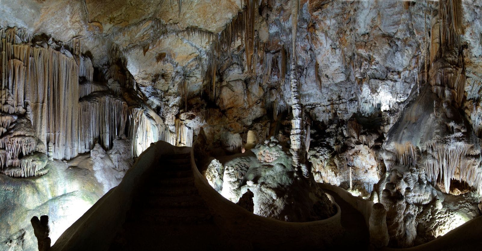 Interior de cova poc il·luminada amb estalactites i estalagmites, passarel·la il·luminada i escales de pedra que condueixen a un nivell superior.