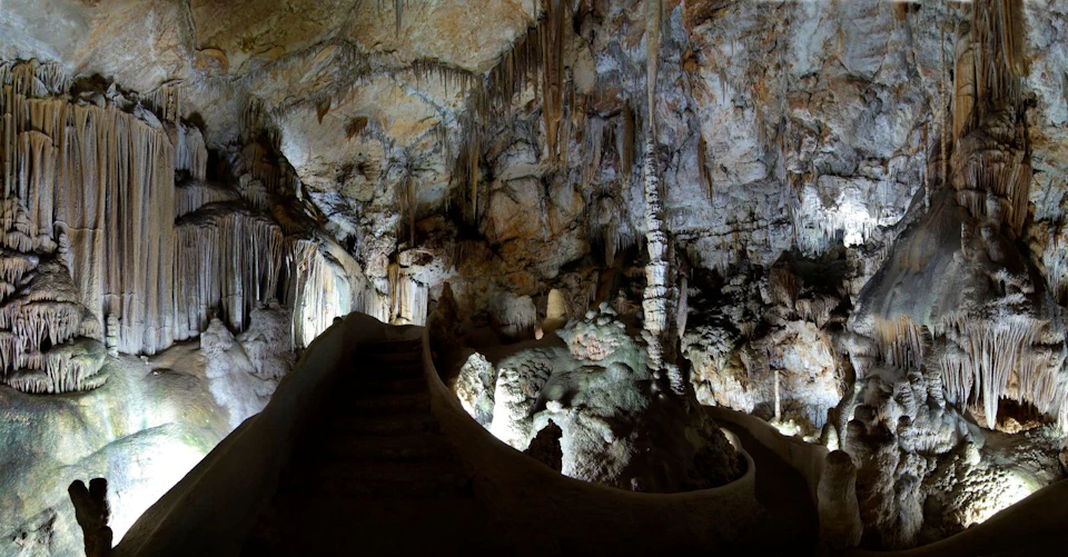 Dimly lit cave interior with stalactites and stalagmites, illuminated walkway, and stone steps leading to an upper level.
