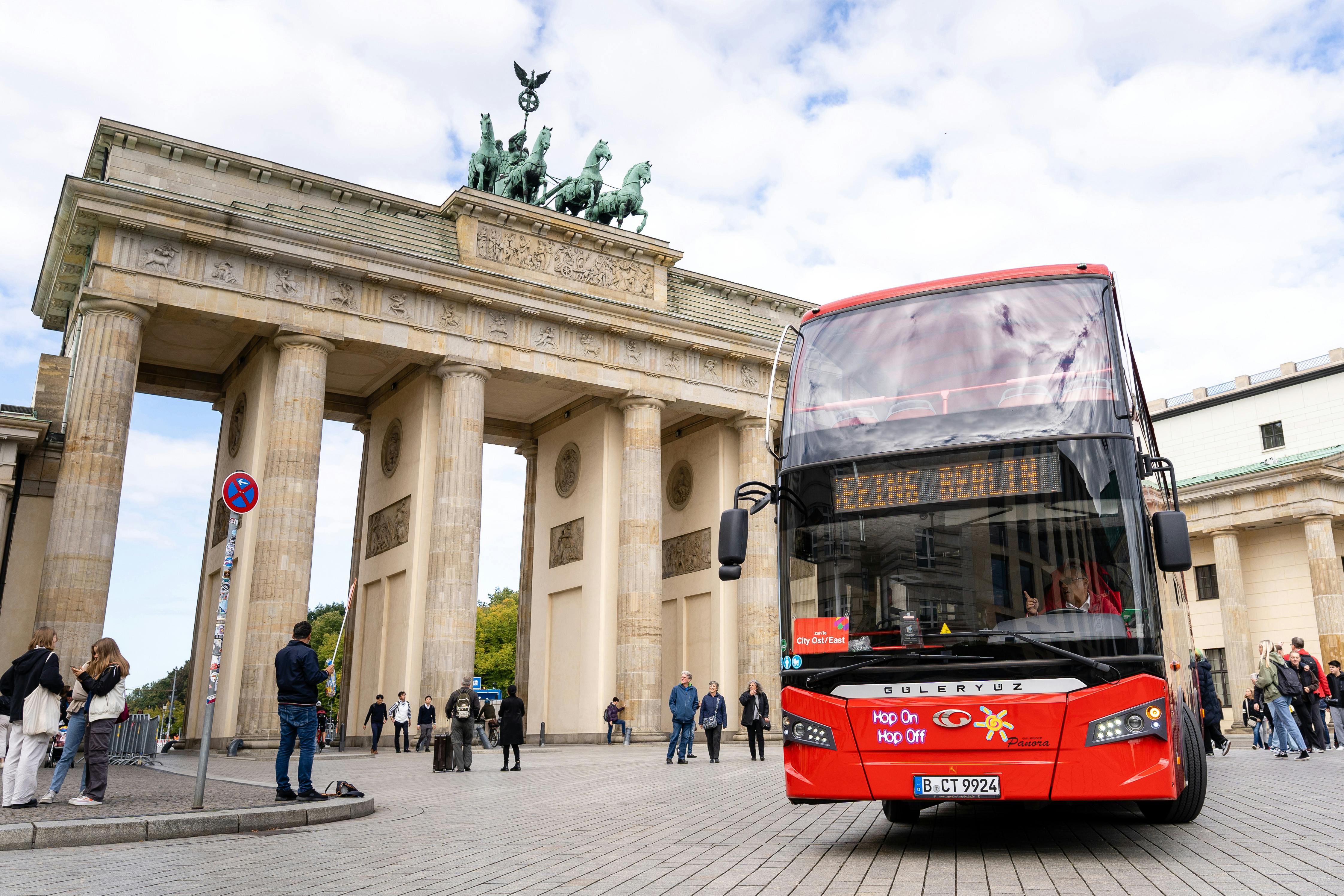 Un bus touristique rouge à impériale devant la porte de Brandebourg, avec des gens qui marchent et prennent des photos.
