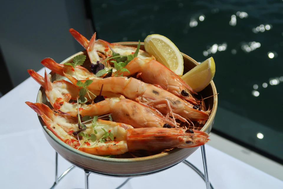A bowl of grilled prawns garnished with herbs and lemon slices, set on a white tablecloth, with water visible in the background.