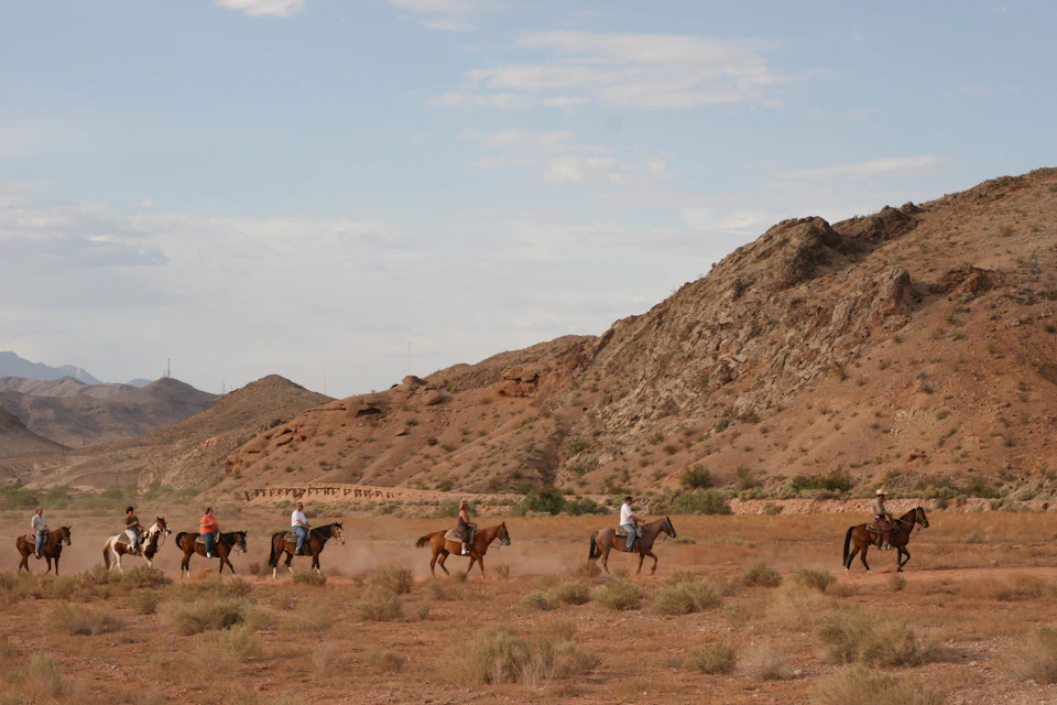 Four people riding horses on a dry, grassy landscape with rocky hills and a partly cloudy sky in the background.