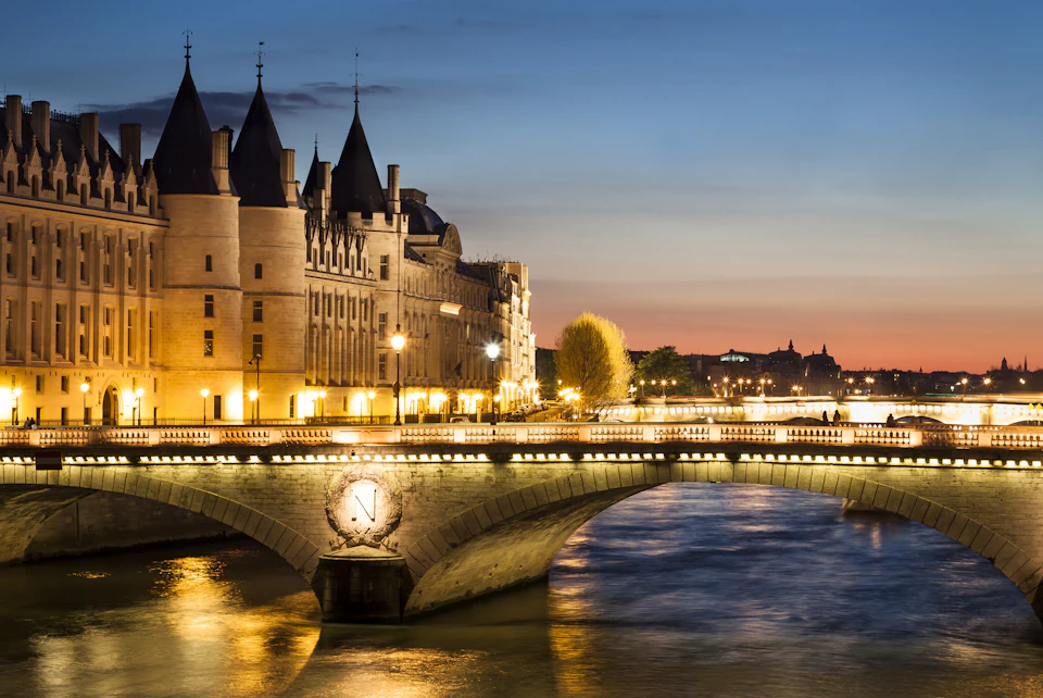 Historic building with spires by a river, illuminated bridge, and twilight sky. Quiet cityscape with lights reflecting on water.