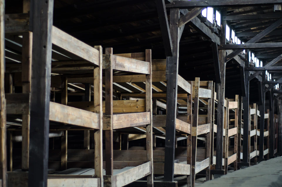 A dimly lit room with rows of wooden bunk beds, supported by dark wooden beams, and small windows at the top.