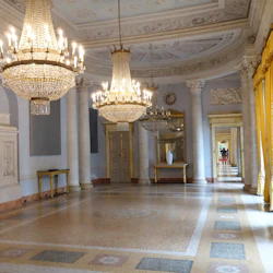 En elegant hall med utsmyckade ljuskronor, pelare, gula gardiner, speglar och dekorativa bord under högt dekorativt tak.