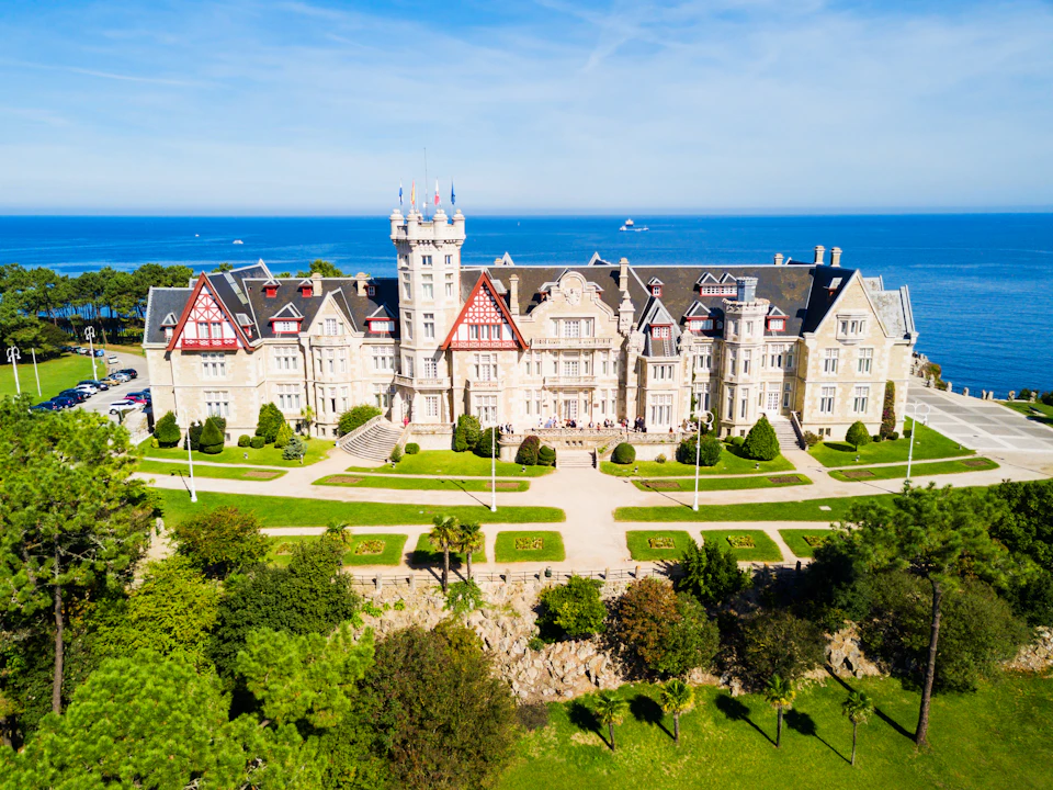Aerial view of a grand, historic building with a central tower, surrounded by gardens, facing the sea under a blue sky.