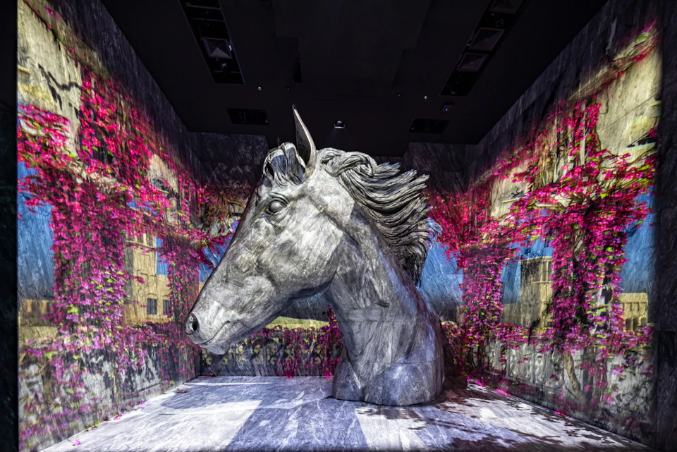 A large sculpted horse head displayed in an indoor exhibit, surrounded by colorful projected visuals on the walls.