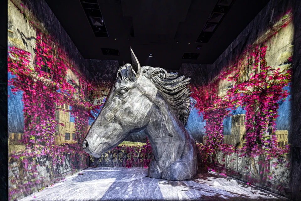 A large sculpted horse head displayed in an indoor exhibit, surrounded by colorful projected visuals on the walls.