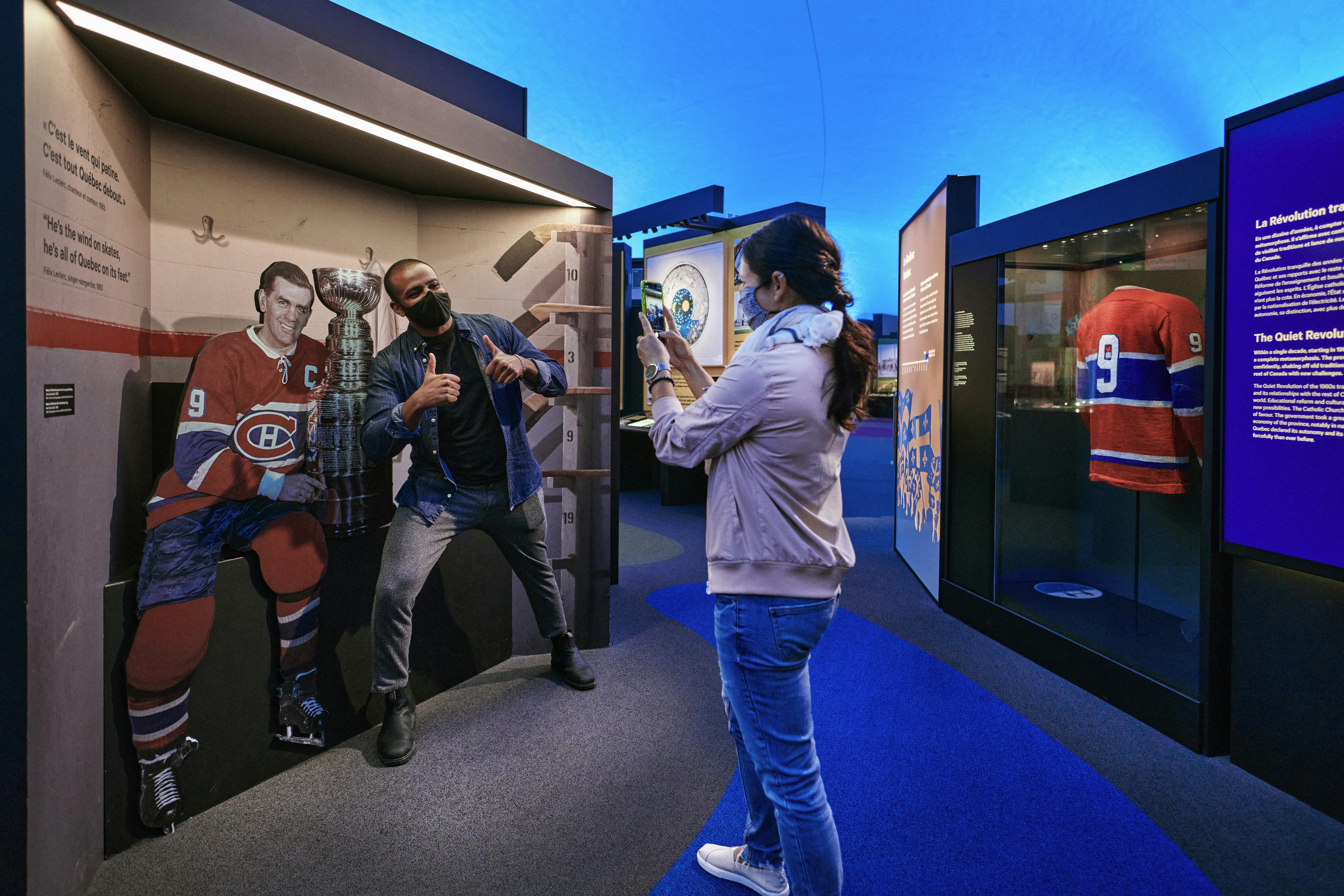 一个人在曲棍球运动员和斯坦利杯复制品旁边摆姿势，另一个人用智能手机拍照。