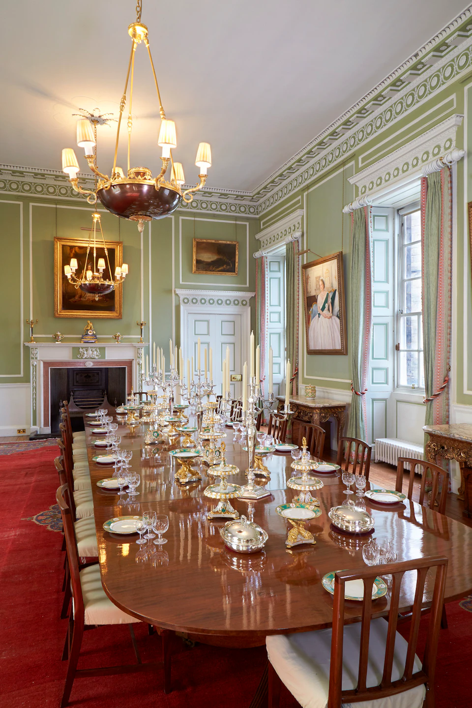 An elegant dining room with a long wooden table, set with silverware and candlesticks. Chandeliers, paintings, and tall windows adorn the space.